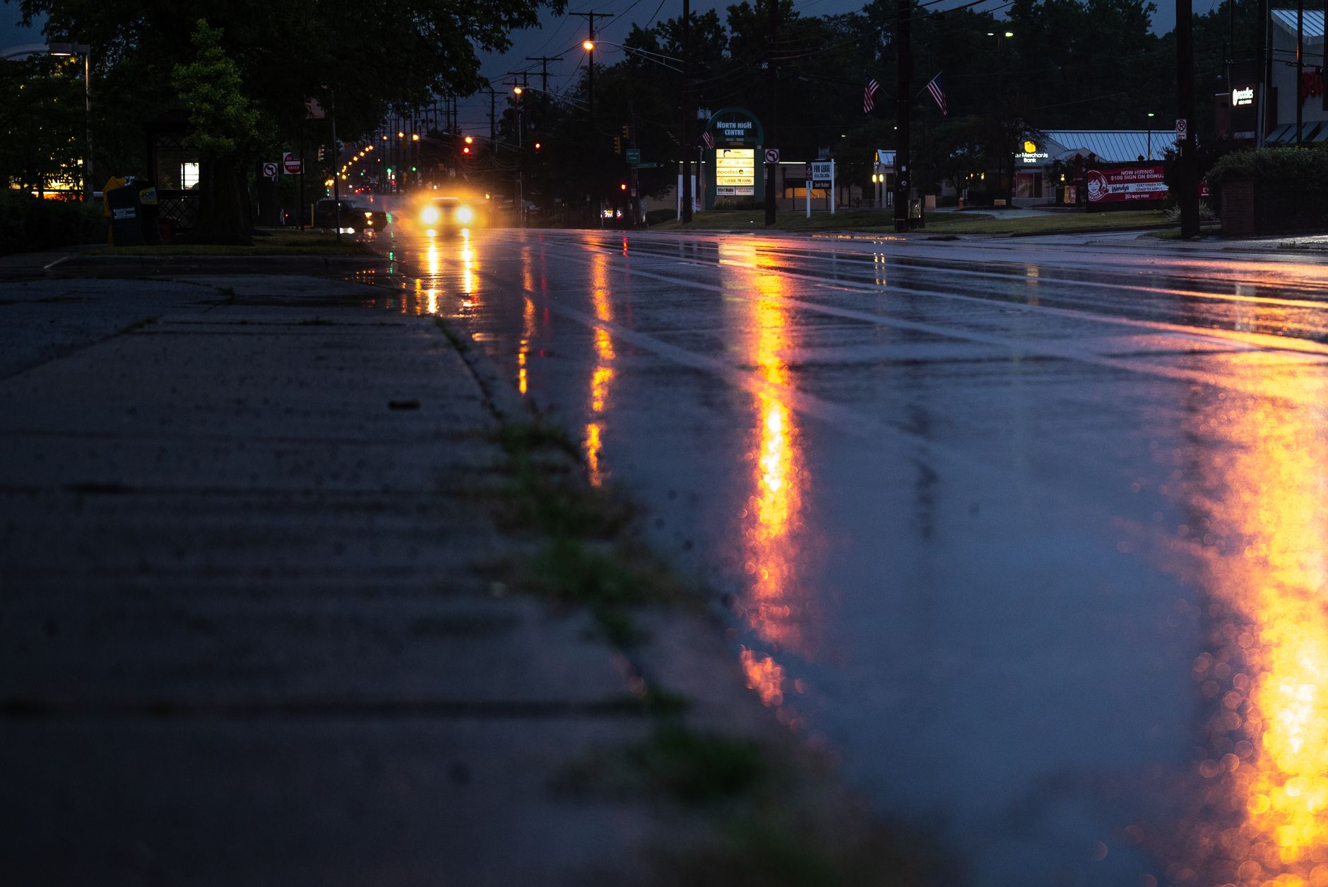 A rain-soaked town street at night reflecting headlights and traffic signals, colours smeared across wet tarmac